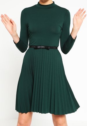 Frau trägt ein dunkelgrünes, langärmeliges, plissiertes Kleid mit schwarzem Gürtel und hat die Hände neben den Schultern erhoben, vor weißem Hintergrund.