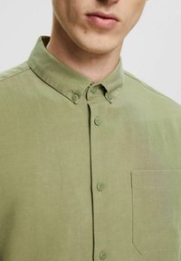 Camicia verde chiaro con bottoni davanti, colletto, texture sottile, taschino sul petto e bottoni abbinati, che mostra tessuto di qualità e design su misura.