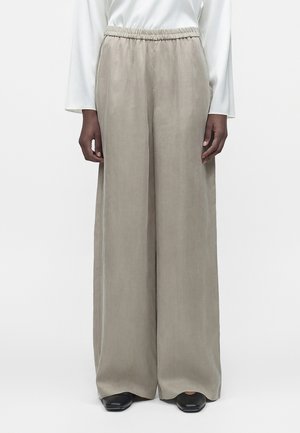 Personne portant un pantalon beige à jambes larges avec taille élastique, un haut blanc à manches longues et des chaussures noires, debout sur un fond uni.