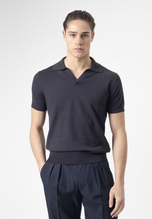 Joven con el cabello peinado hacia atrás, que lleva una camiseta polo ajustada de manga corta en oscuro y pantalones plisados oscuros, de pie con las manos en los bolsillos.