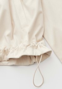 Manche de veste beige avec un poignet élastiqué et une boucle à cordon réglable sur un fond blanc uni.