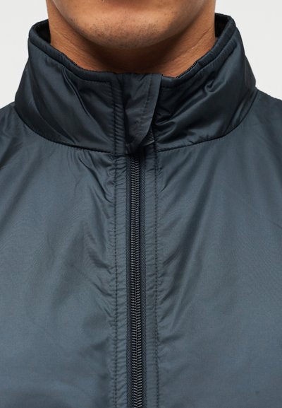 Σκούρο μπλε, ελαφρύ υφασμάτινο μπουφάν με ψηλό γιακά, φερμουάρ ολόκληρο μήκος και λείο ύφασμα. Χωρίς ορατά σχέδια ή διακοσμητικά στοιχεία.