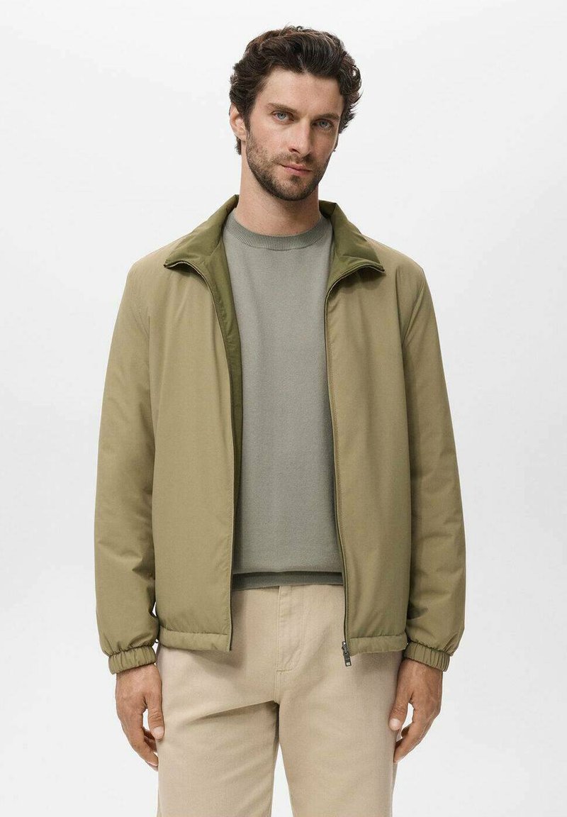 Mann mit kurzen lockigen Haaren, der eine khakifarbene Zip-Up-Jacke, einen hellgrauen Pullover und beige Hosen vor einem schlichten weißen Hintergrund trägt.