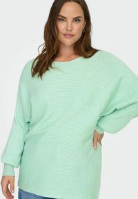 Vaaleanvihreä ribbineulottu ylipitkä neule pyöreällä pääntiellä, lasketuilla hartioilla ja pitkillä hihoilla, yhdistettynä sinisiin farkkuihin.