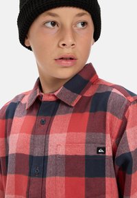 Rood en marineblauw geruit flanellen overhemd met een button-down kraag, voorzien van een borstzak en een klein logo-label aan de zijkant. Gedragen met een zwarte gebreide muts.