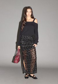 Maglione nero lavorato a maglia con design scollato sulle spalle, abbinato a una gonna nera trasparente con motivo texturizzato. Un borsa bordeaux completa il look.