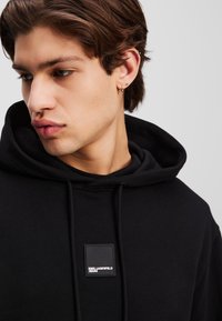 Čierna mikina s kapucňou a textúrovanou tkaninou, s predným vreckom, šnúrkou na kapucni a obdĺžnikovou nášivkou na hrudi.