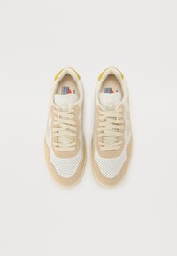 Sneakers beige e bianchi con texture perforata, lacci e un accento giallo sul tallone. Logo impresso sul lato e sulla linguetta.
