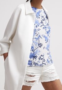 Camisa blanca de manga larga sobre una blusa azul con estampado floral, combinada con pantalones cortos blancos rasgados. Texturas similares al algodón son visibles en todas las prendas.
