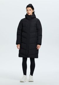 Sort puffet frakke med hætte, der har quiltet design, lange ærmer og en mid-længde. Parret med sorte leggings og hvide sneakers.
