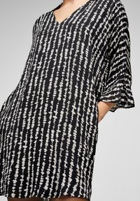 Svart och vit mönstrad tunika med V-ringning, korta volangärmar och lös passform; har en vertikal design med oregelbundna prickar och linjer.