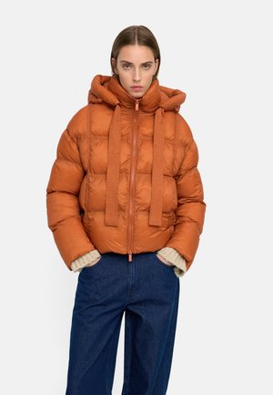 Kobieta ubrana w pomarańczową kurtkę puchową z kapturem i dużymi ściągaczami, w połączeniu z niebieskimi dżinsami i beżowymi dzianinowymi mankietami.