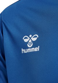 Blå sportskjorte med glat stof, der har et hvidt logo fra mærket "hummel" og et karakteristisk designelement, der ligner vinger.