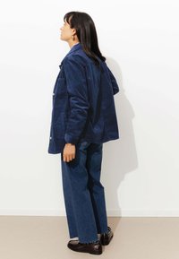 Veste en velours côtelé bleu marine à coupe décontractée, avec poche poitrine et boutons argentés, associée à un pantalon en denim ample et des chaussures noires brillantes.