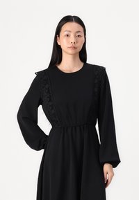 Black long-sleeve dress featuring lace detailing on the shoulders, a round neckline, and an elasticized waist for a relaxed fit.
