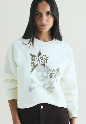 Donna con capelli lunghi e scuri che indossa una felpa bianca con una grafica di teschio, ossa incrociate e stelle con scritto "Rock Spirit."