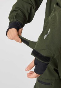 Grön utomhusjacka med texturerat tyg, kardborrefästen vid ärmslut och svarta innerärmar. Har en logga och en fungerande dragkedjeficka.