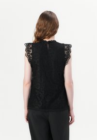 Top in pizzo nero con design a maniche scallop, modelli floreali intricati e dettaglio a chiave sul retro, abbinato a pantaloni neri.