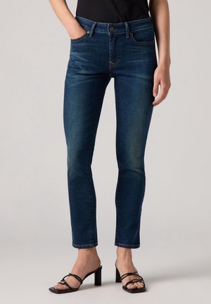 Personne portant un jean slim bleu foncé et des sandales à talons noires debout devant un fond uni.
