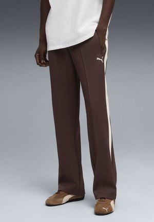 Mees, kes kannab pruune Puma-vabaaja pükse valgete küljeribadega, pruune Puma tossusid ja valget T-särki, seisab hallil põrandal.