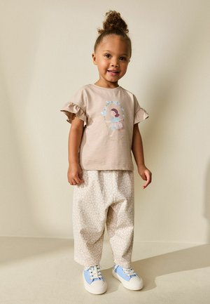 Gaiši bēša īsroku krekls ar volānu malām un ziedu rakstu, kopā ar vaļējām krēmkrāsas biksēm ar smalku rakstu un zilām sporta kurpēm.