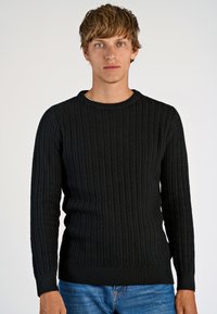Maglione nero lavorato a maglia con un design a costine testurizzate, collo a giro, e maniche lunghe, abbinato a jeans blu.