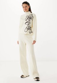 Maglione avorio con ricami floreali neri, colletto alto e spalle scese, abbinato a pantaloni larghi avorio e sneakers con dettagli neri.