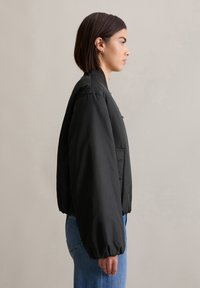 Zwarte bomberjack met een hoge kraag, elastische manchetten en een losse pasvorm, gecombineerd met lichtblauwe jeans. Glad textuur met een minimalistisch ontwerp.