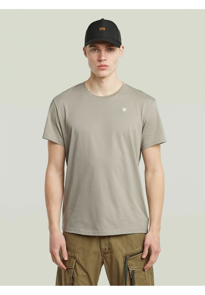 Beżowa bawełniana koszulka z krótkim rękawem i okrągłym dekoltem, z małym logo na lewej piersi. Noszona z brązowymi spodniami cargo i czapką w kolorze czarnym.