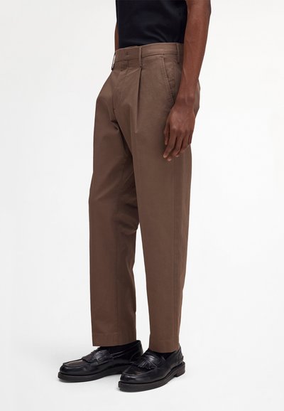 Homme portant un pantalon plissé marron et des mocassins noirs, debout devant un fond clair uni.