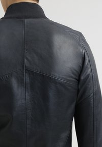 Svart läderjacka med texturerad yta, med en ribbad krage och kantiga sömmar på ryggen. Slät, figursydd silhuett.