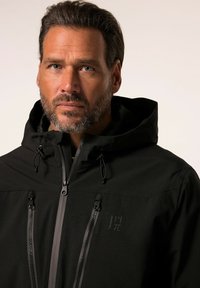 Schwarze wasserdichte Jacke mit Kapuze, ausgestattet mit mehreren Reißverschluss-Taschen und Logodetail auf der Brust. Glatte, matte Textur.