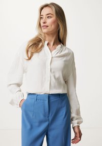 Camicia bianca a maniche lunghe con chiusura a bottoni e scollo raccolto, abbinata a pantaloni blu con vestibilità sartoriale e tasche laterali.