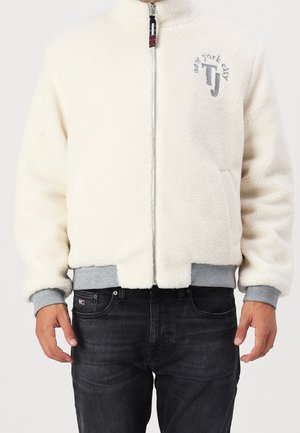 Mann trägt eine weiße Fleecejacke mit Reißverschluss, grauen Bündchen und Saum, mit „New York City TJ“ auf der Brust bestickt, und schwarze Jeans.