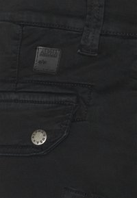 Crne traperice imaju kvadratnu aplikaciju s "ALPHA INDUSTRIES" logotipom, srebrne detalje i džep sa zatvaranjem na dugme.