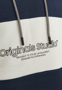 Sweat-shirt bleu marine et crème avec un logo sur le devant "Originals Studio", présentant un tissu texturé et un cordon de serrage contrasté avec des embouts noirs.