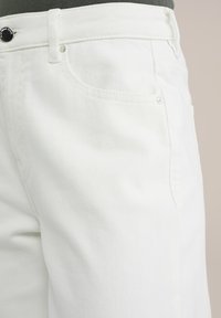 Vita denimjeans med texturerad yta, framfickor och knäppning, med en närbild som betonar tygets detaljer.