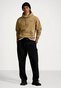 Φούτερ με κουκούλα σε μπεζ απόχρωση, με τσέπη μπροστά και κορδόνι, φορεμένο πάνω από λευκό μπλουζάκι; συνδυασμένο με μαύρο cargo παντελόνι και μαύρα αθλητικά παπούτσια.