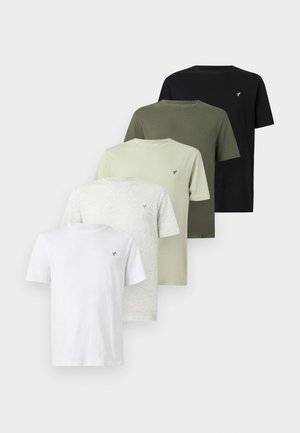 Homme en chemise noire à manches courtes, joggeurs gris clair avec cordon de serrage et baskets blanches, debout les mains dans les poches devant un fond blanc.
