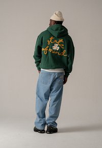Zelená mikina s kapucňou so žltým vyšitým nápisom „state of mind“, v kombinácii so svetlomodrými voľnými džínsami a čiernymi topánkami. Na hlave béžová čiapka.