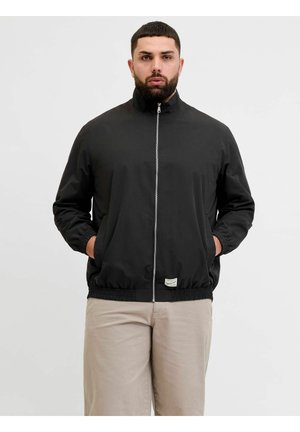 Uomo che indossa una giacca nera con cerniera, mani nelle tasche e pantaloni beige, in piedi davanti a uno sfondo chiaro e uniforme.