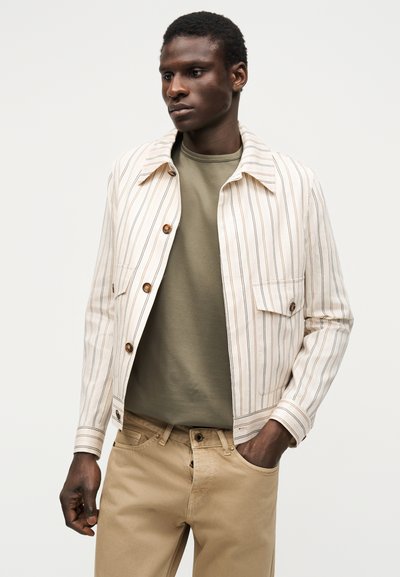 Mees kannab beežikat triibulist jopet oliivrohelise särgi ja helepruunide pükstega, seisab ühe käega taskus, neutraalne taust.