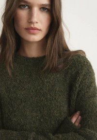 Pulover verde măsliniu dintr-un material texturat, cu un guler rotund și mâneci lungi, având un aspect moale și pufos.
