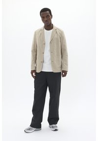 Beige linen blazer with two front pockets and button closure, worn over a white T-shirt. Paired with black trousers and light gray sneakers.