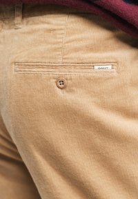 Helle beige Cord-Hosen mit vertikaler Rippstruktur, ausgestattet mit einer Gesäßtasche mit Knopf und einem kleinen weißen Etikett mit der Aufschrift "GANT."