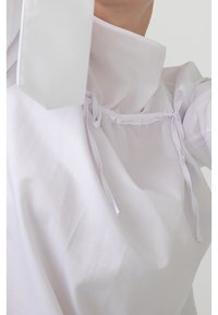 Witte blouse met een hoge kraag, voorzien van een geplooide voorkant met koordjes. Zachte, gladde stof en een ontspannen pasvorm creëren een minimalistisch ontwerp.