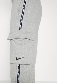 Šedé tepláky s cargo kapsou, s černým logem Nike a bočním pruhem s opakujícím se textem "Nike" v černé a bílé barvě.