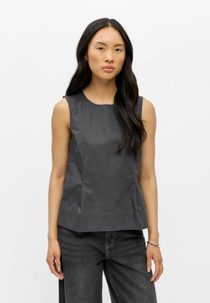 Femme aux longs cheveux noirs portant un haut gris foncé sans manches et un jean noir sur un fond blanc uni.