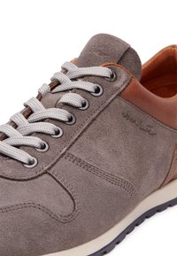 Szare zamszowe sneakersy z brązowymi skórzanymi akcentami, szarymi sznurowadłami, okrągłymi metalowymi oczkami i gumową podeszwą z teksturowanym gripem.