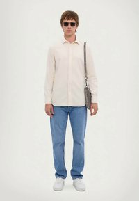 Modelis vilki šviesiai grietinėlės spalvos marškinėlius su mygtukais, mėlynus džinsus ir baltus sportbačius, laiko pilką per petį nešiojamą krepšį, dėvi tamsius saulės akinius.